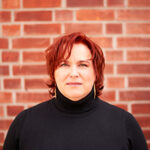Eine Frau mit rot gefärbten Haaren und im schwarzen Rollkragenpullover steht vor einer Ziegelwand und blickt direkt in die Kamera.