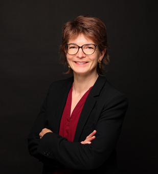 Eine Frau mit Brille, roter Bluse und schwarzer Jacke lächelt freundlich.