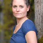 Eine Frau mit lockigen, zusammengebundenen Haaren lehnt an einem Baum und schaut in die Kamera.