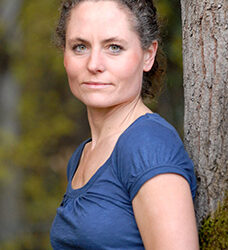 Eine Frau mit lockigen, zusammengebundenen Haaren lehnt an einem Baum und schaut in die Kamera.