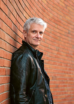 Ein Mann mit grauen Haaren und Lederjacke lehnt an einer roten Ziegelwand.